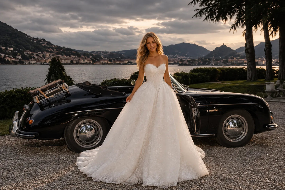 Modern Bride wearing an off-white lace wedding gown beside a vintage convertible overlooking a lake at sunset